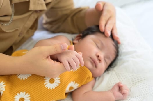 Hand of mother holding finger of newborn hand. Adorable newborn baby sleeping on bed at home while mom lying down nursing baby girl and gently touching on head with love. Care, love and protection concept.