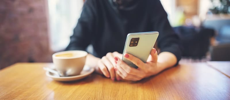 Woman holding phone in her hands and sitting in cafe at the table with cup of coffee. Concept of the break after work and leisure.