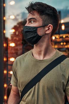 Young man standing at store front in the city center in the evening, looking away, wearing the face mask to avoid virus infection and to prevent the spread of disease in time of coronavirus