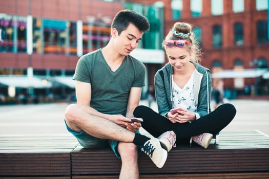 Couple of friends, teenage girl and boy,  having fun together with smartphones,  sitting in center of town, spending time together