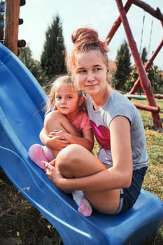 Teenage girl taking a photo her younger sister in a home playground in a backyard. Happy smiling sisters having fun on a slide together on summer day. Real people, authentic situations