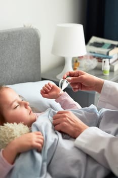 Doctor visiting little patient at home. Measuring the temperature of sick girl lying in bed. Woman wearing uniform and face mask. Medical treatment
