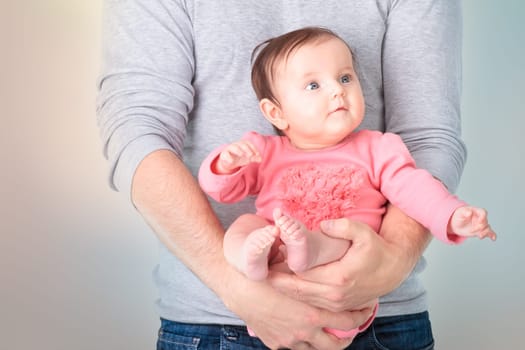 Baby sitting on fathers hands. Baby is wearing pink body suit and looking at right side