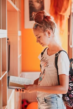 Teenage girl looking for books in local library. Young woman with a backpack standing by a bookshelf indoors. Real people, authentic situations
