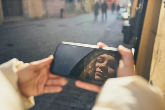 Young woman having video call, talking remotely, taking selfie photo holding smartphone. Face reflected in screen of smartphone