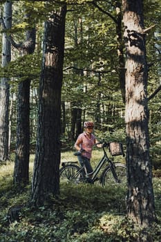 Active woman spending free summer vacation time on a bicycle trip in a forest. Woman wearing bicycle helmet and gloves holding bike with basket standing behind the trees