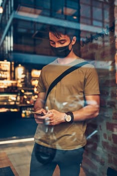 Young man standing in coffee shop at store front in the city center in the evening, wearing the face mask to avoid virus infection and to prevent the spread of disease in time of coronavirus