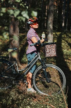 Active woman spending free summer vacation time on a bicycle trip in a forest. Woman wearing bicycle helmet and gloves holding bike with basket and drinking a water from bottle