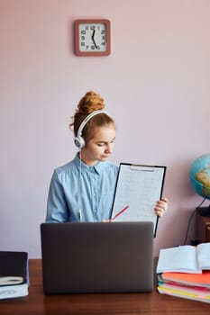 Young woman student showing homework, having classes, learning online, watching lesson remotely, listening to professor, talking with classmates on video call from home during quarantine. Young girl using laptop, headphones, books, manuals sitting at a desk
