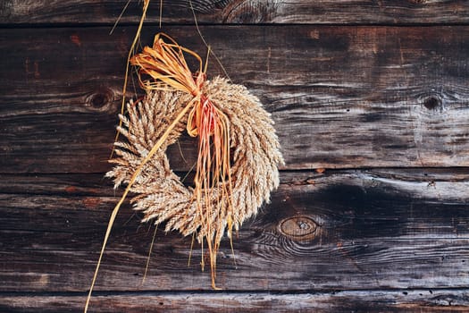 Wreath of golden ears of wheat, dried flowers and herbs tied with red ribbon hung on wooden wall. Natural rustic decoration