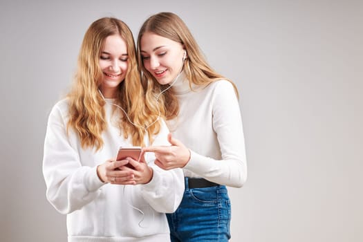 Young women girls listening to music together streaming content having fun watching video enjoying video chat talking with friends making gestures faces using smartphone earphones headphones standing over plain grey background