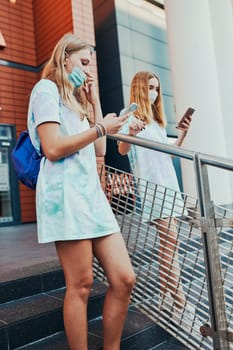 Young women girls spending time together downtown using smartphones social media sending texts watching photos and videos while walking in the city center. Girls wearing the face masks to avoid virus infection and to prevent the spread of disease in time of coronavirus