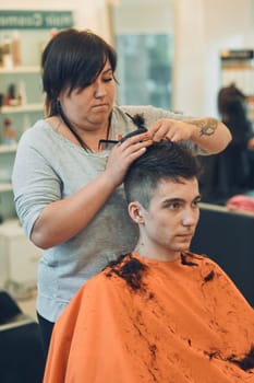 Hairdresser haircutting styling young man's hair in barbershop. Young woman working as a hairdresser in hair salon. Real people, authentic situations
