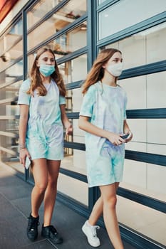 Young women girls spending time together downtown walking along store front in the city center. Girls wearing the face masks to avoid virus infection and to prevent the spread of disease in time of coronavirus
