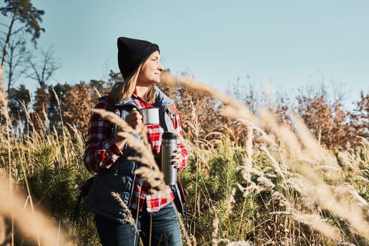 Woman taking break and relaxing with cup of coffee during summer trip. Woman standing on trail and looking away. Female with backpack hiking through tall grass along path in mountains. Spending summer vacation close to nature