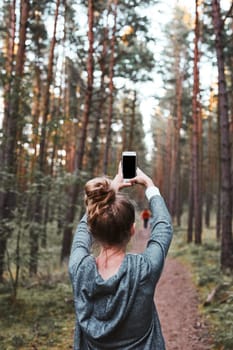 Young woman taking photos using smartphone during walk in a forest on summer vacation trip. Back view of girl holding smartphone