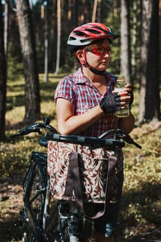 Active woman spending free summer vacation time on a bicycle trip in a forest. Woman wearing bicycle helmet and gloves holding bike with basket and drinking a water from bottle