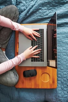Teenage girl working on school tasks remotely on her laptop from home during COVID-19 quarantine. Online course lessons. Learning remotely. Girl sitting on bed in front of computer. View from above