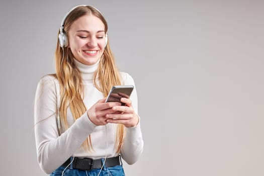 Young woman girl listening to music streaming content having fun watching video enjoying video chat talking with friends making gestures faces using smartphone earphones headphones standing over plain grey background