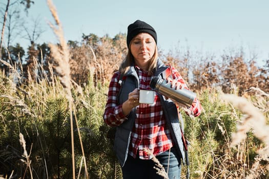 Woman taking break and relaxing with cup of coffee during summer trip. Woman standing on trail and looking away. Female with backpack hiking through tall grass along path in mountains. Spending summer vacation close to nature