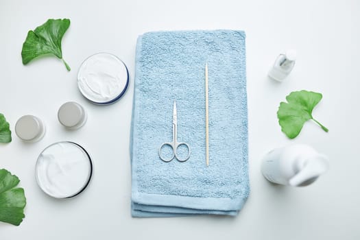 Cosmetic products set, green leaves, blue towel on white table. Spa, manicure, skin care concept. Flat lay, overhead view