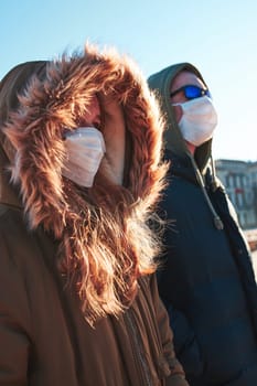 Young woman and man in the hoods wearing the face masks to avoid virus infection and to prevent the spread of disease. People walking in city centre. Virus infection protection. Real people, authentic situations