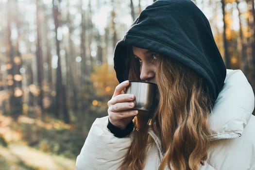 Woman in a hood having break during autumn trip holding cup with hot drink from thermos flask on autumn cold day. Active girl wandering in a forest actively spending time