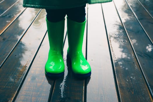 Closeup of legs of kid standing on a porch wearing green wellies and raincoat. Rain boots in bright green color