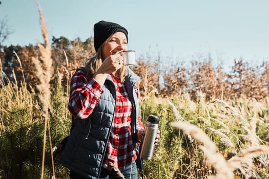 Woman taking break and relaxing with cup of coffee during summer trip. Woman standing on trail and looking away. Woman with backpack hiking through tall grass along path in mountains. Spending summer vacation close to nature