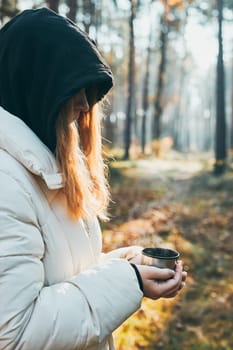 Woman in a hood having break during autumn trip holding cup with hot drink from thermos flask on autumn cold day. Active girl wandering in a forest actively spending time