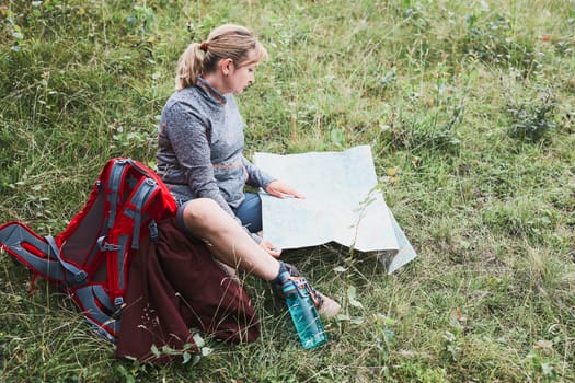 Woman with backpack having break during trip in mountains looking at map sitting on grass on summer vacation day