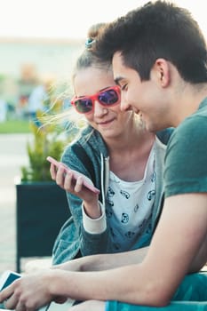 Couple of friends, teenage girl and boy,  having fun using smartphones sitting in center of town, spending time together