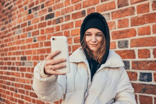 Young woman having video call, talking remotely, taking selfie photo holding smartphone standing at brick wall