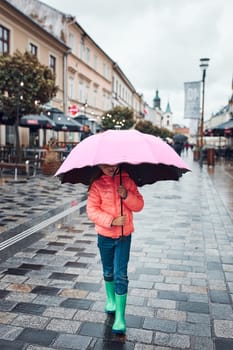 Child holding big pink umbrella walking in a downtown on rainy gloomy autumn day