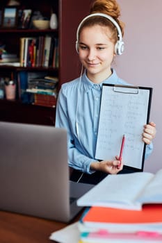 Young woman student showing homework, having classes, learning online, watching lesson remotely, listening to professor, talking with classmates on video call from home during quarantine. Young girl using laptop, headphones, books, manuals sitting at a desk
