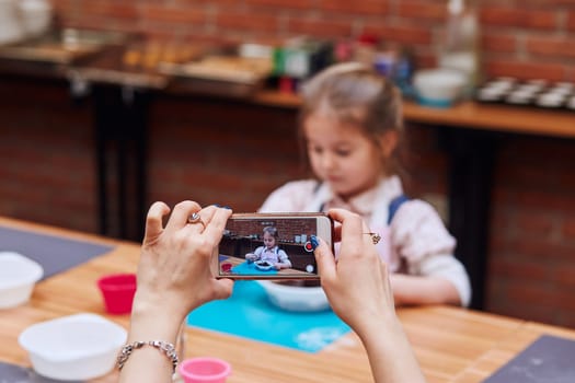 Mom recording her little daughter while taking part in baking workshop. Baking classes for children,  aspiring little chefs. Learning to cook. Combining and stirring prepared ingredients. Real people, authentic situations