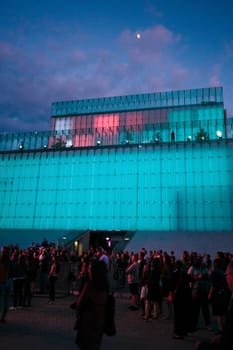 Festival event party concert with people standing on a square in a city center at night. Backlit glass construction above them