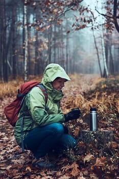 Woman with backpack having break during autumn trip drinking a hot drink from thermos flask on autumn cold day. Active middle age woman wandering around a forest actively spending time