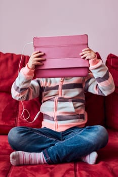 Little girl preschooler learning online solving puzzles playing educational games listetning to music and sounds using tablet and headpphones sitting on sofa at home