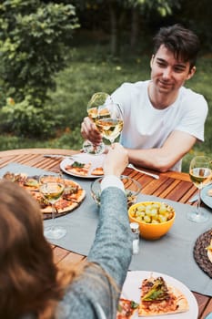 Friends making toast during summer picnic outdoor dinner in a home garden. Close up of people holding wine glasses with white wine over table with pizza, salads and fruits. Dinner in a orchard in a backyard