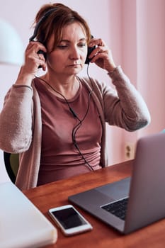 Woman working talking doing her job remotely during video chat phone call on laptop from home. Woman sitting at desk in front of computer looking at screen using headphones and smartphone. Concept of remote working