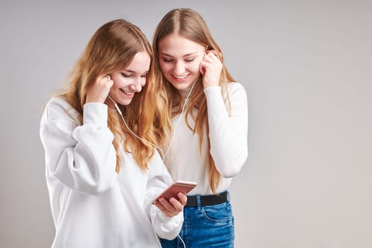 Young women girls listening to music together streaming content having fun watching video enjoying video chat talking with friends making gestures faces using smartphone earphones headphones standing over plain grey background