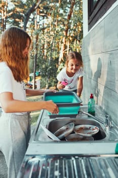 Teenager girl washing up the dishes pots and plates with help her younger sister in the outdoor kitchen during vacations on camping. Camp life. Sisters spending time and working together