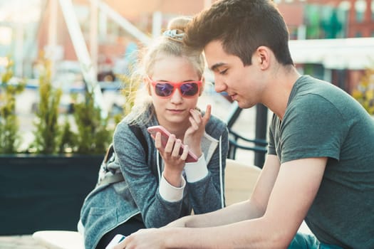 Couple of friends, teenage girl and boy,  having fun using smartphones sitting in center of town, spending time together