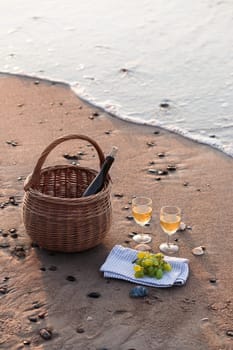 Two wine glasses with white wine standing on sand, on beach, beside grapes and wicker basket with bottle of wine