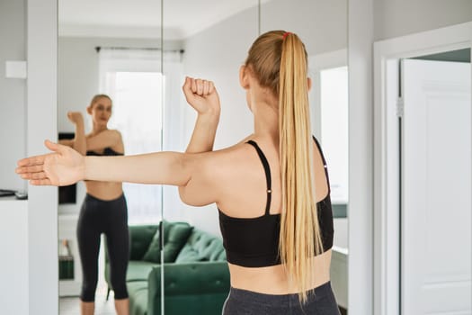 Young woman in sportswear stretches arms in her living room in front of a mirror, back view