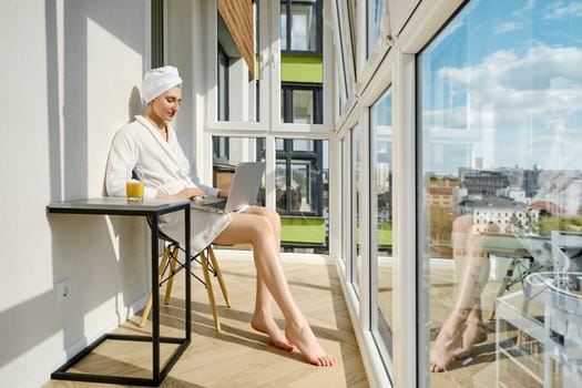 Barefoot woman in a bathrobe sits on balcony of her new high-rise apartment and using laptop