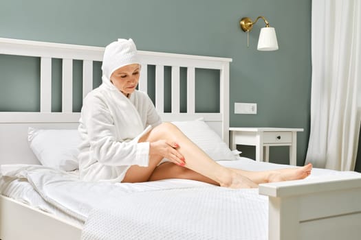 Senior woman sitting on bed and applying lotion on her feet after shower