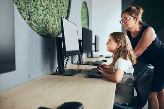 Teacher assisting schoolgirl while computer class at primary school. Child learning computer on elementary computer science class. Teacher helping to find task solution. Giving clues. Smart little girl learning online at school. Back to school