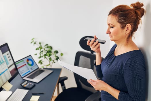 Woman entrepreneur having business  conversation on mobile phone. Businesswoman working in office holding documents and smartphone. Woman standing by desk with computers